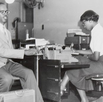Scheltes collection archival greyscale photograph of a Black blind man with a briefcase, sitting beside a desk. A Black woman is sitting at the desk, looking down and writing.
                  