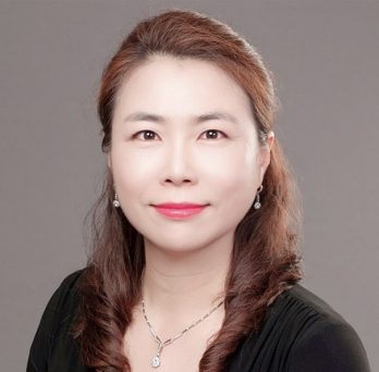Portrait of Jung Mi Scoulas who is wearing black blouse and necklace and earrings in front of a neutral background.
                  