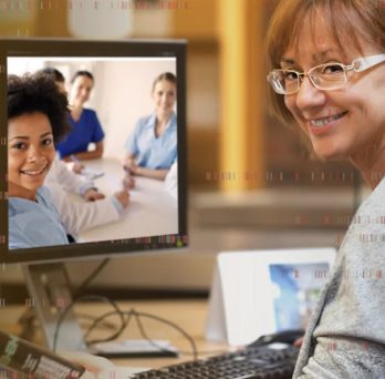 Woman with glasses smiling looking over her shoulder while sitting at a desk with a computer. Computer shows image three people smiling.
                  