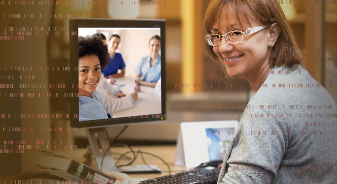 Woman with glasses smiling looking over her shoulder while sitting at a desk with a computer. Computer shows image three people smiling.