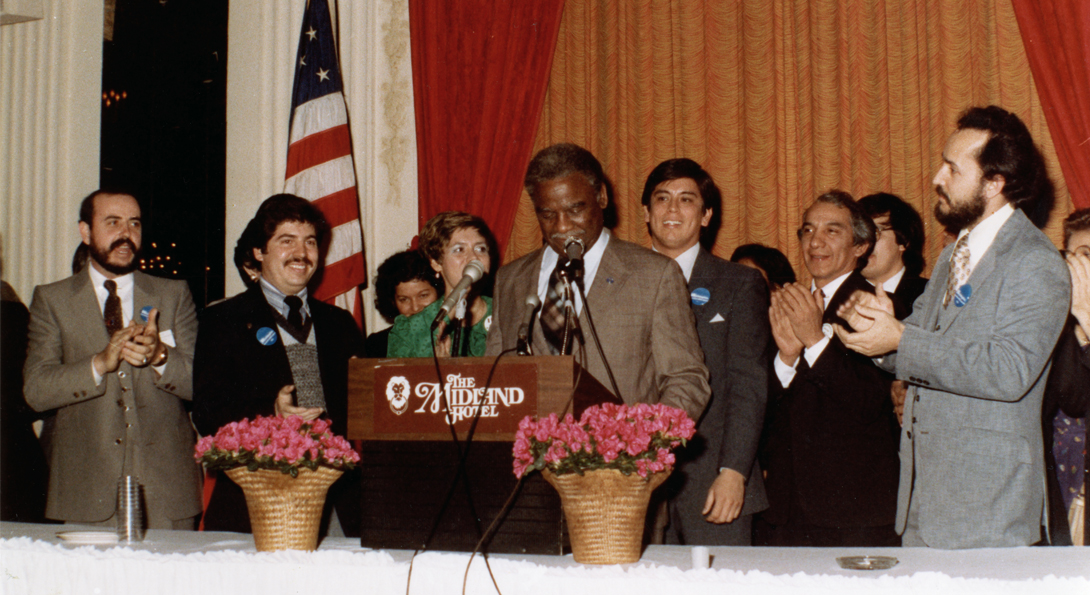 Group of people, including Rudy Lozano, in business attire surrounding and clapping for the late Chicago Mayor Harold Washington who is standing at a podium speaking.