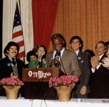 Group of people, including Rudy Lozano, in business attire surrounding and clapping for the late Chicago Mayor Harold Washington who is standing at a podium speaking. 