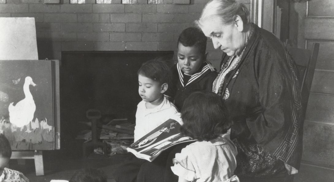 Archival black and white photograph of Jane Addams sitting in a chair reading a book to children who are gathered around her chair. there is a painting of a duck on an ease and a fireplace in the background.