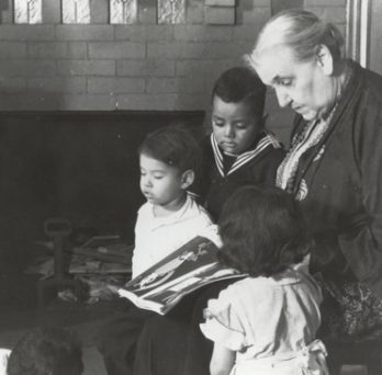 Archival black and white photograph of Jane Addams sitting in a chair reading a book to children who are gathered around her chair. there is a painting of a duck on an ease and a fireplace in the background. 