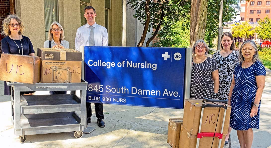 Grant stakeholders standing around the College of Nursing sign outside of the building with 3D printer and supplies on carts.