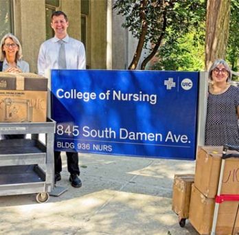 Grant stakeholders standing around the College of Nursing sign outside of the building with 3D printer and supplies on carts. 