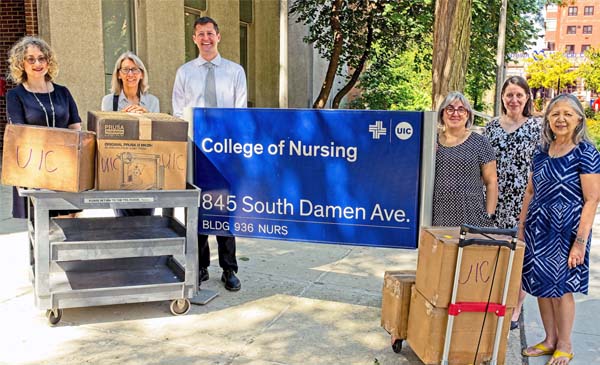 Grant stakeholders standing around the College of Nursing sign outside of the building with 3D printer and supplies on carts.