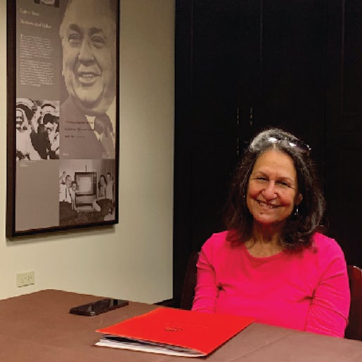 Portrait of Helen Shiller in pink shirt sitting at table with a red folder. A photo montage of images of Richard J. Daley hangs in the background.