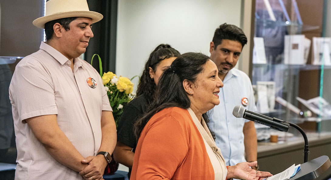 Profile view of Lupe Lozano speaking into a microphone at a podium surrounded by her two sons and granddaughter in the Circle Reading Room.
