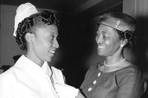 Undated historical greyscale photograph of a Black nurse and another Black woman smiling at each other, celebrating the nurse's graduation. The nurse is wearing a white nursing uniform. The other woman is wearing a matching hat and dress and white gloves and a pearl necklace.