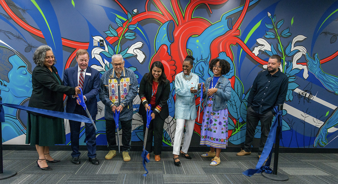 Ribbon cutting ceremony for the Odeh Health Equity Center. Group of people laughing and smiling as they cut a ribbon in front of a richly colored blue, red and green mural featuring a human heart, figures and flowers.