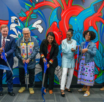 Ribbon cutting ceremony for the Odeh Health Equity Center. Group of people laughing and smiling as they cut a ribbon in front of a richly colored blue red and green mural featuring a human heart, figures and flowers. 