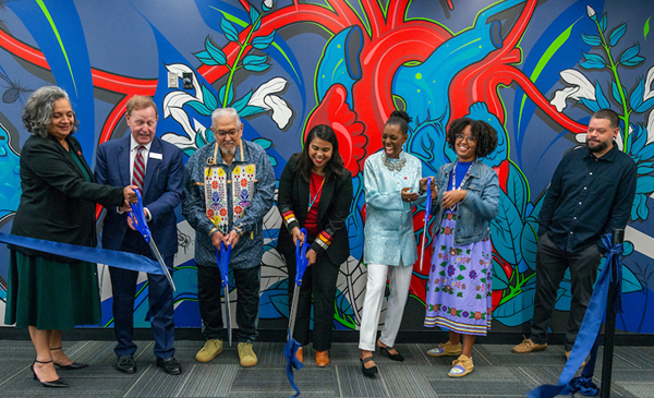 Ribbon cutting ceremony for the Odeh Health Equity Center. Group of people laughing and smiling as they cut a ribbon in front of a richly colored blue, red and green mural featuring a human heart, figures and flowers.