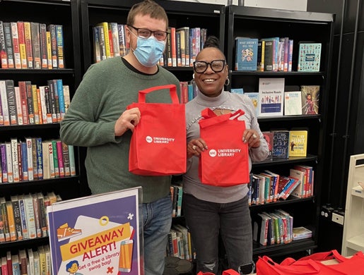 Two Library employees smiling and holding up red swag bags in front of bookshelves.