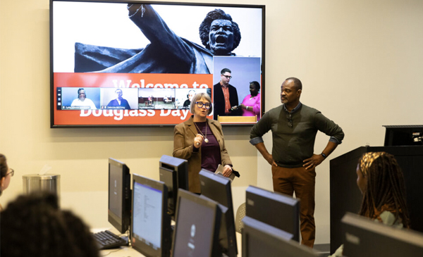 Amy Bailey and Joseph Jewell discuss the Douglass Day transcription event with participants in a computer classroom. People are sitting at the computers. There is a screen behind them that says 