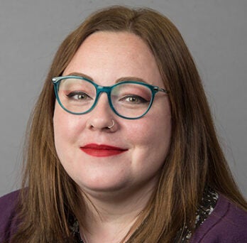 Emily Gilbert wearing teal glasses and a purple sweater on a neutral background. 