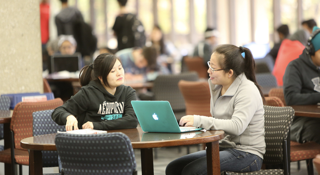 Two students studying at a table with a computer at the library. Many other students sit and stand in the background.
