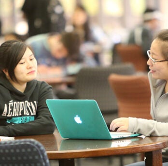 Two students studying at a table with a computer at the library. Many other students sit and stand in the background. 