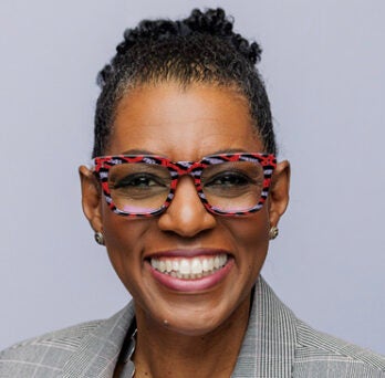 Portrait of Rhea Ballard-Thrower smiling in grey suit, red, black and grey flecked glasses. 