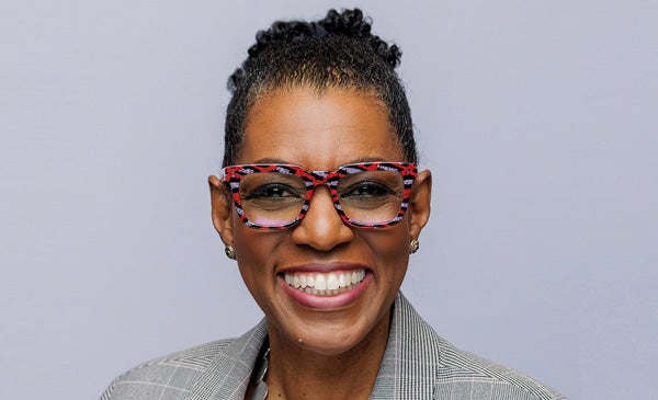 Portrait of Rhea Ballard-Thrower, smiling and wearing a grey suit and red, black and grey flected glasses.