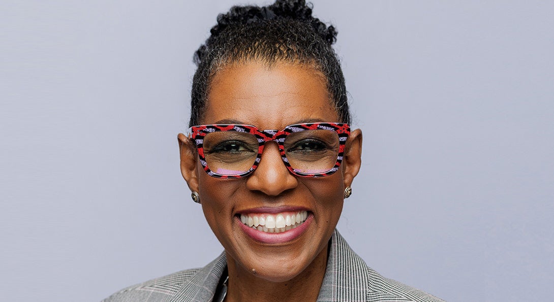 Portrait of Rhea Ballard-Thrower, smiling and wearing a grey suit and red, black and grey flected glasses.