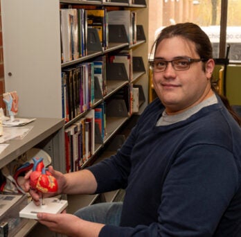 David Johnson holding an anatomical model of a human heart with shelves of books in the background at the Library of the Health Sciences-Chicago 