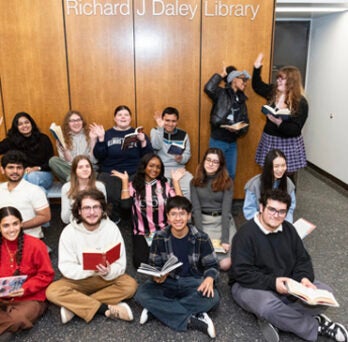 Group of Library Student Employees, some seated on the floor, some standing with books. Some are waving and some are making silly faces and smiling. Standing students are giving each other a high five. 