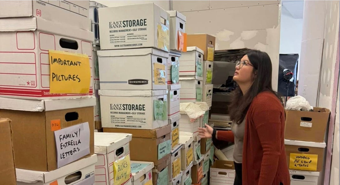 Grace deVega standing in front of towering stacks of boxes of archival materials. Two boxes are labeled 'Important Pictures' and 'Family Estrella Letters'
