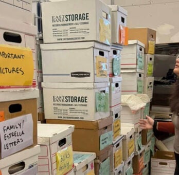 Grace deVega standing in front of towering stacks of boxes of archival materials. Two boxes are labeled 'Important Pictures' and 'Family Estrella Letters' 